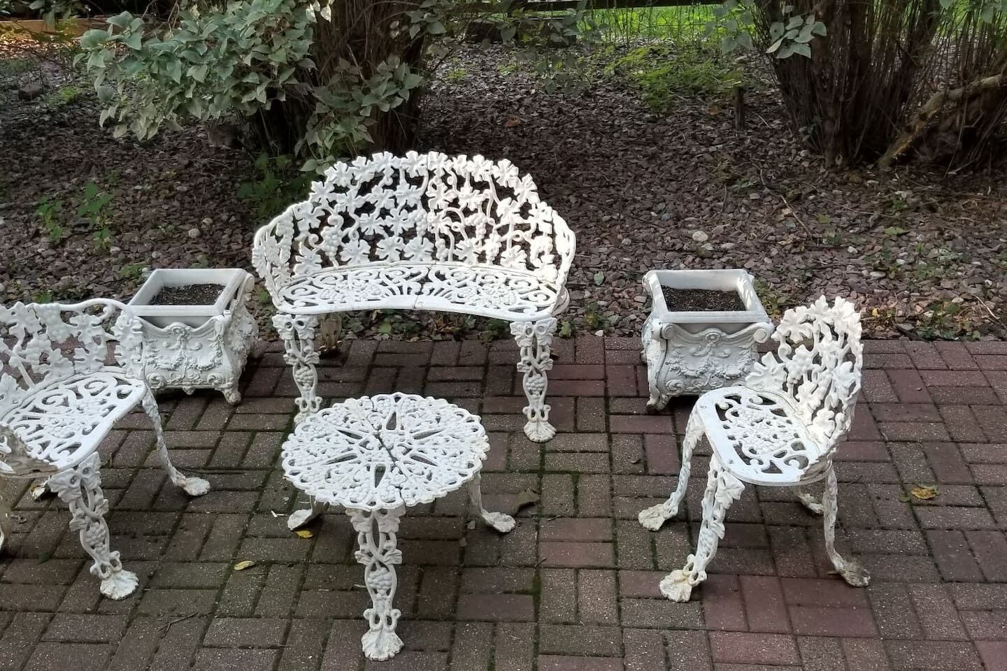 A set of ornate white metal outdoor furniture consisting of a table, two chairs, and a bench, arranged on a paved surface with a natural backdrop.