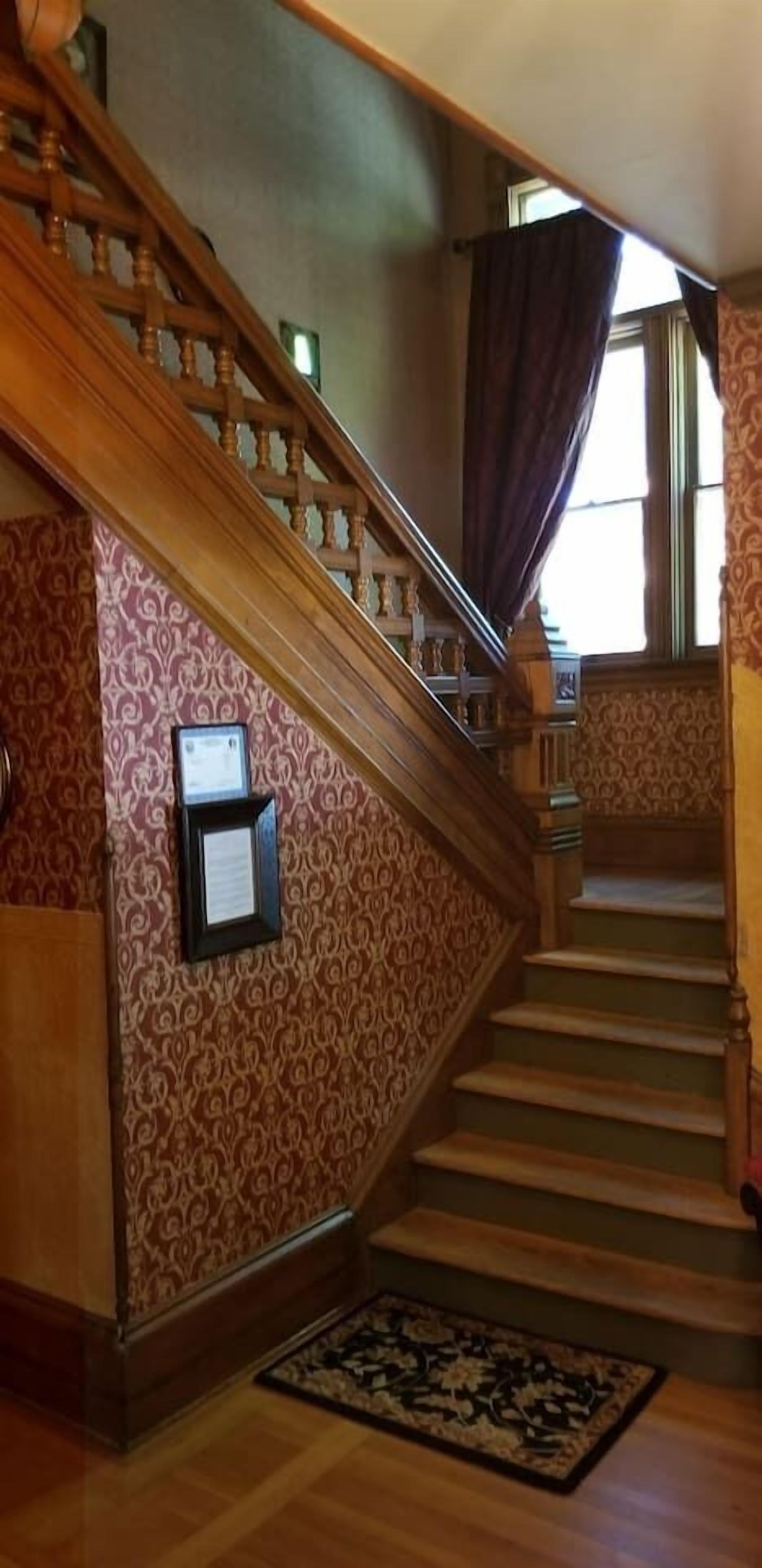 A grand staircase with ornate woodwork and patterned wallpaper, leading to a landing with a framed notice.