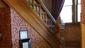 Una imponente escalera con detalles de madera decorativos y un papel tapiz estampado, que conduce a un rellano con un aviso enmarcado.