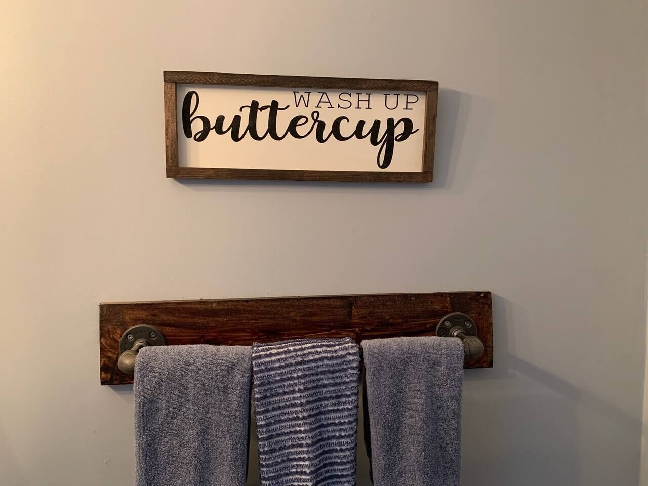 A wall-mounted towel rack with two towels and a sign above it that reads 'WASH UP buttercup.'
