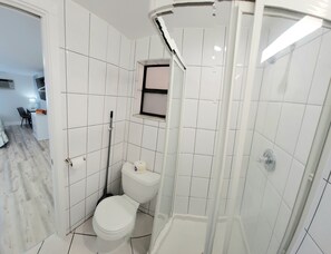 A compact bathroom with white tiles, a toilet, a shower, and a small window. - Satellite Motel - Sea & Bay View Bungalow 127 (Treasure Island)