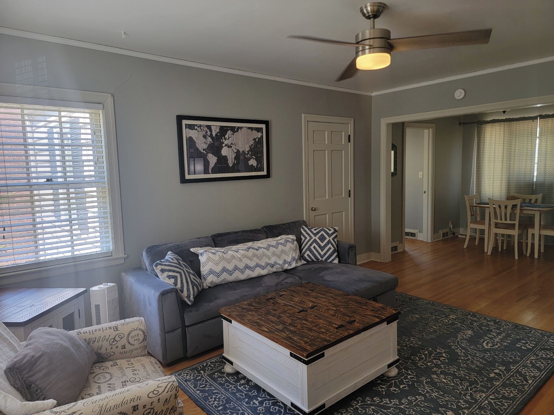 A living room with a gray sofa, a wooden coffee table, a world map on the wall, and a dining area with a table and chairs.