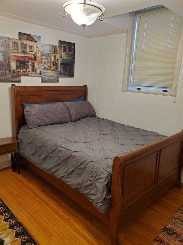 A bedroom with a wooden bed, a nightstand, a window with blinds, and three paintings on the wall.