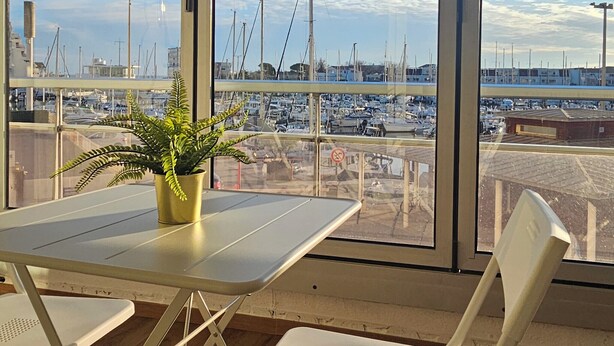 Habitación con un amplio ventanal que da a un puerto deportivo con barcos, una planta en maceta sobre una mesa y una silla.