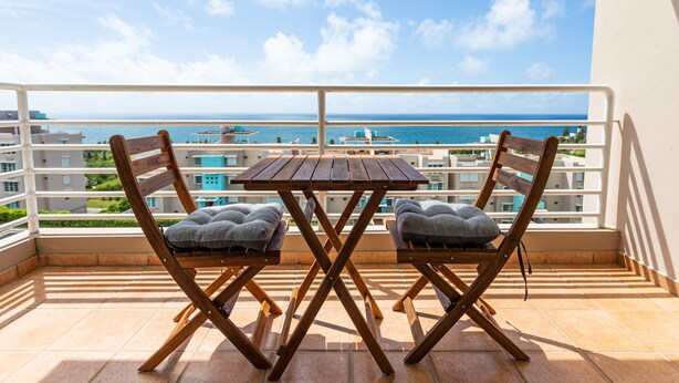 Uma varanda com uma mesa de madeira e duas cadeiras acolchoadas, com vista para a costa e edifícios ao fundo.