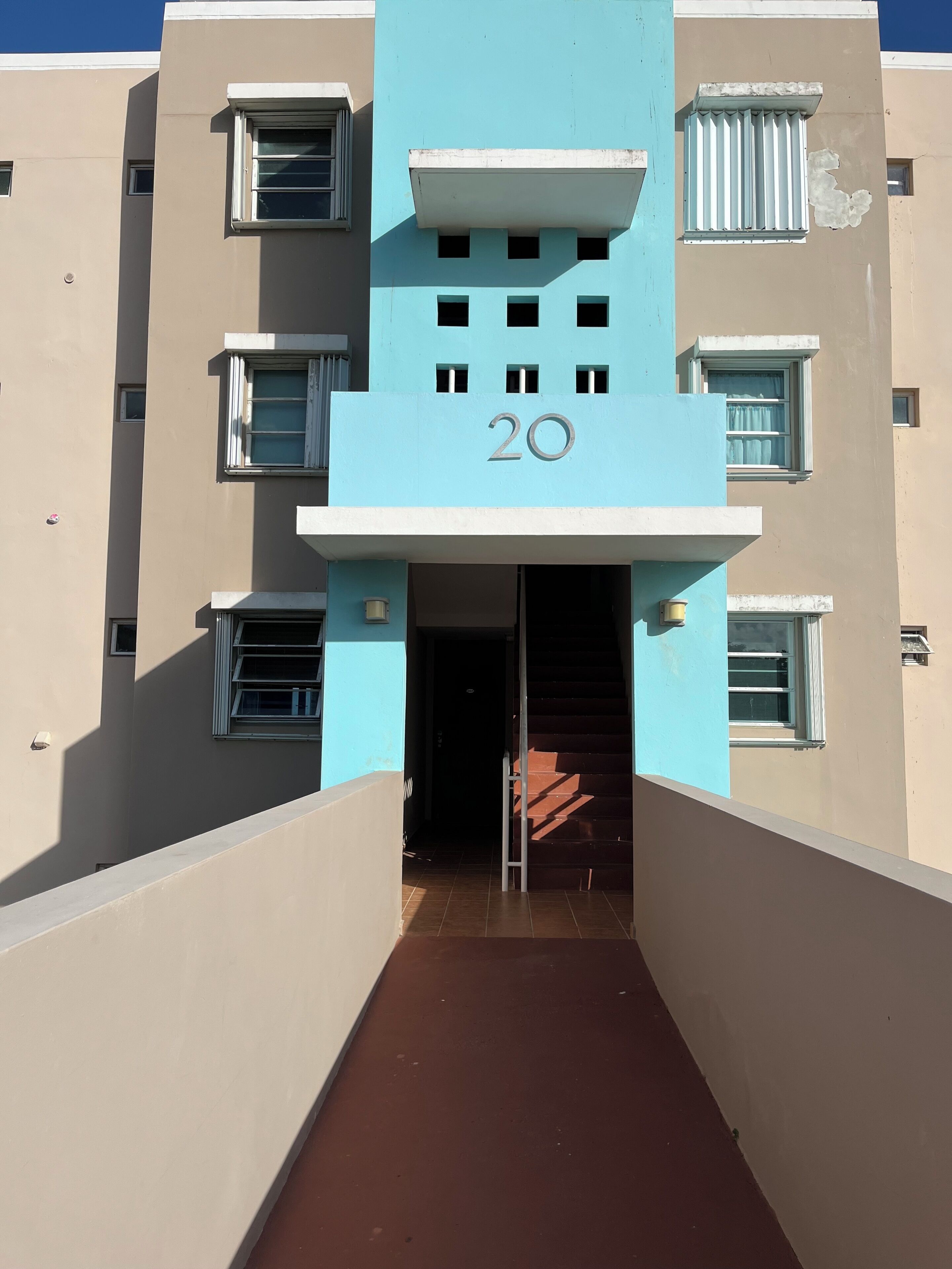 Un edificio con un número '20' destacado en la fachada, una entrada central con escaleras y varias ventanas con persianas.