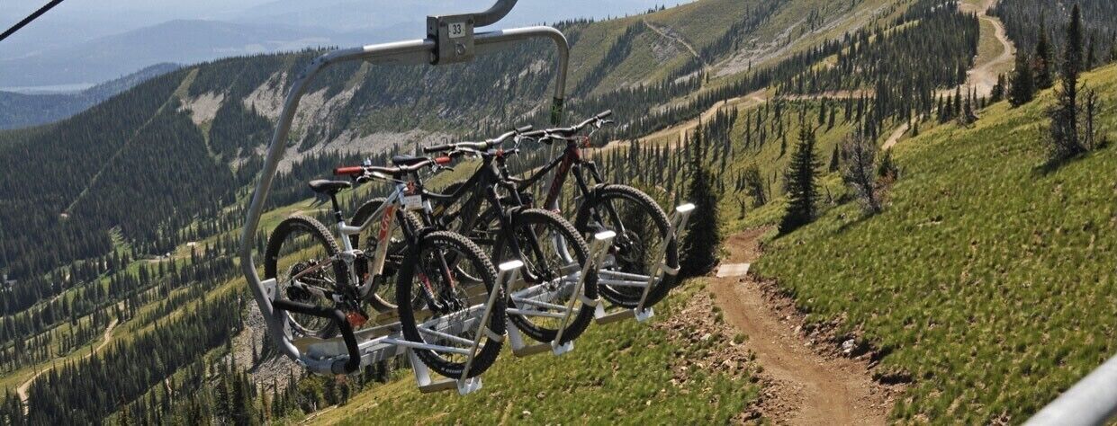 A mountain bike rack with several bikes parked on it, overlooking a forested valley with a dirt path.