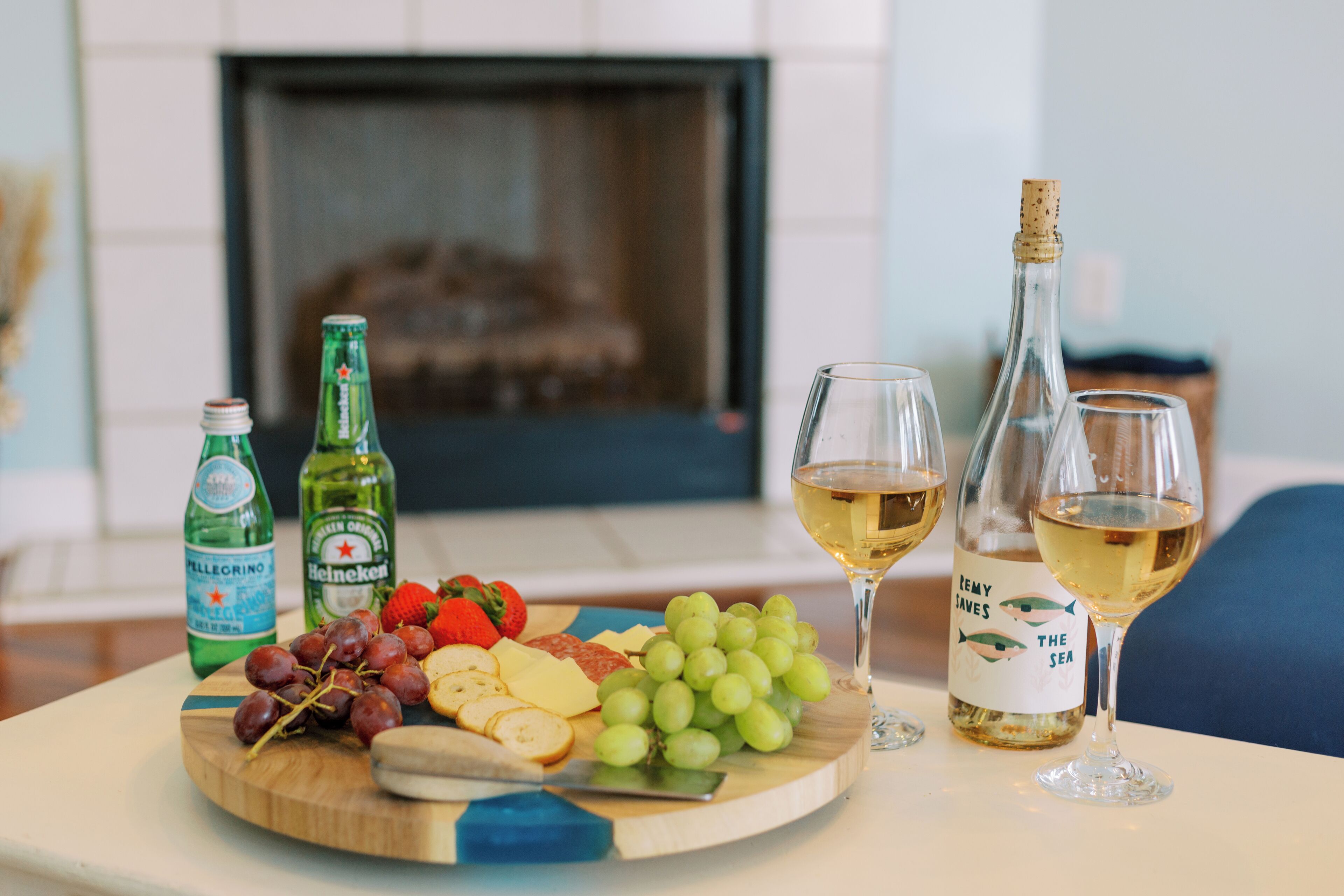 A wooden board with cheese, grapes, and sliced bread, accompanied by two bottles of beer and two glasses of white wine, set on a table in a room with a fireplace in the background.