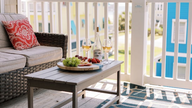 Mesa de madeira com um prato de frutas e copos de vinho em um alpendre com guarda de proteção branca.