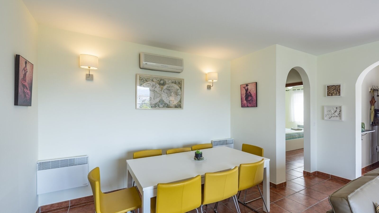 A dining area with a white table and yellow chairs, a world map on the wall, and a small plant on the table.