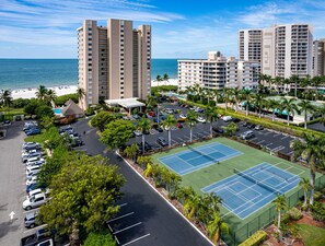 A high-rise hotel with a tennis court and parking lot, overlooking a beach.