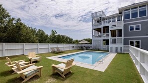 A backyard with a swimming pool, wooden deck chairs, and a multi-level building with balconies. - Great for Families, Salt Water Pool, Sea & Sound Views, Quiet Gated Cul-De-Sac (Kitty Hawk)