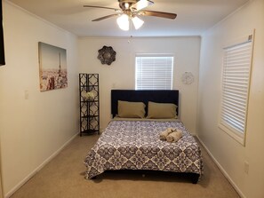 Une chambre à coucher comprenant un lit, un ventilateur de plafond, une fenêtre avec des stores, un élément décoratif mural et un tableau encadré.
