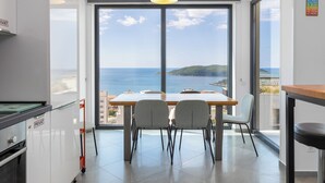 A modern kitchen with a large window overlooking the sea, a dining table with chairs, and a view of a distant island. - Modern Top Floor Sea View 3BR-Pool Spa Gym Luxury (Becici)
