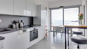 A modern kitchen with white cabinetry, a built-in oven, and a dishwasher. There is a dining area with a table and chairs, and a large window offering a view of the sea. - Modern Top Floor Sea View 3BR-Pool Spa Gym Luxury (Becici)