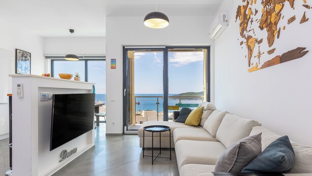 A modern living room with a flat-screen TV, a sofa, and a small table. There is a large window with a view of the sea. - Modern Top Floor Sea View 3BR-Pool Spa Gym Luxury (Becici)