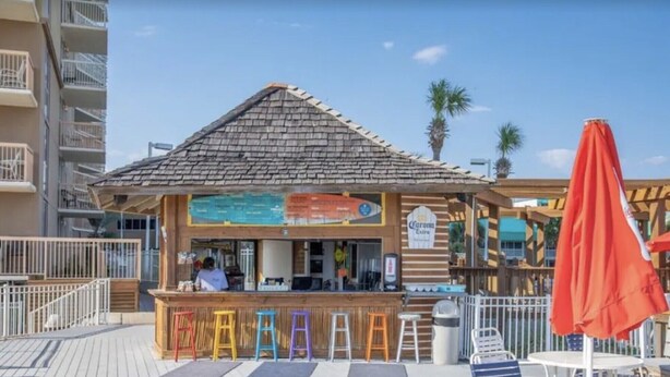 Eine Strandbar mit Holzstruktur, bunten Sitzhocker und einem roten Sonnenschirm.