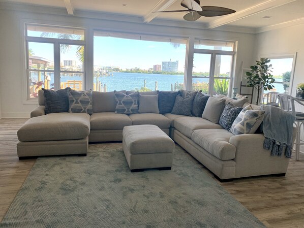 A spacious living room with a large sectional sofa, a ceiling fan, and a view of a body of water through large windows. - The Family Vacation Dream Home - 4BR/4BA, 5 minutes from the beach (Clearwater)