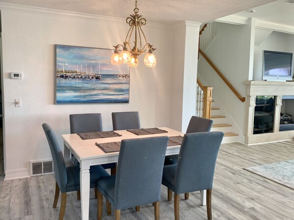 A dining area with a white table, blue chairs, a chandelier, and a painting of a harbor.