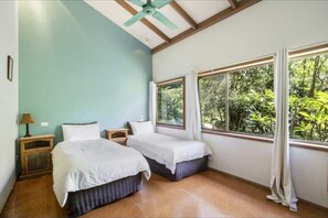 Two single beds with white linens are positioned against a light blue wall, offering a serene view of lush greenery through a large window.