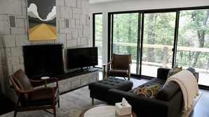 Una sala de estar moderna con chimenea de piedra, un televisor de pantalla plana, sillones de cuero, un sofá de color oscuro y ventanas grandes con vistas a árboles.