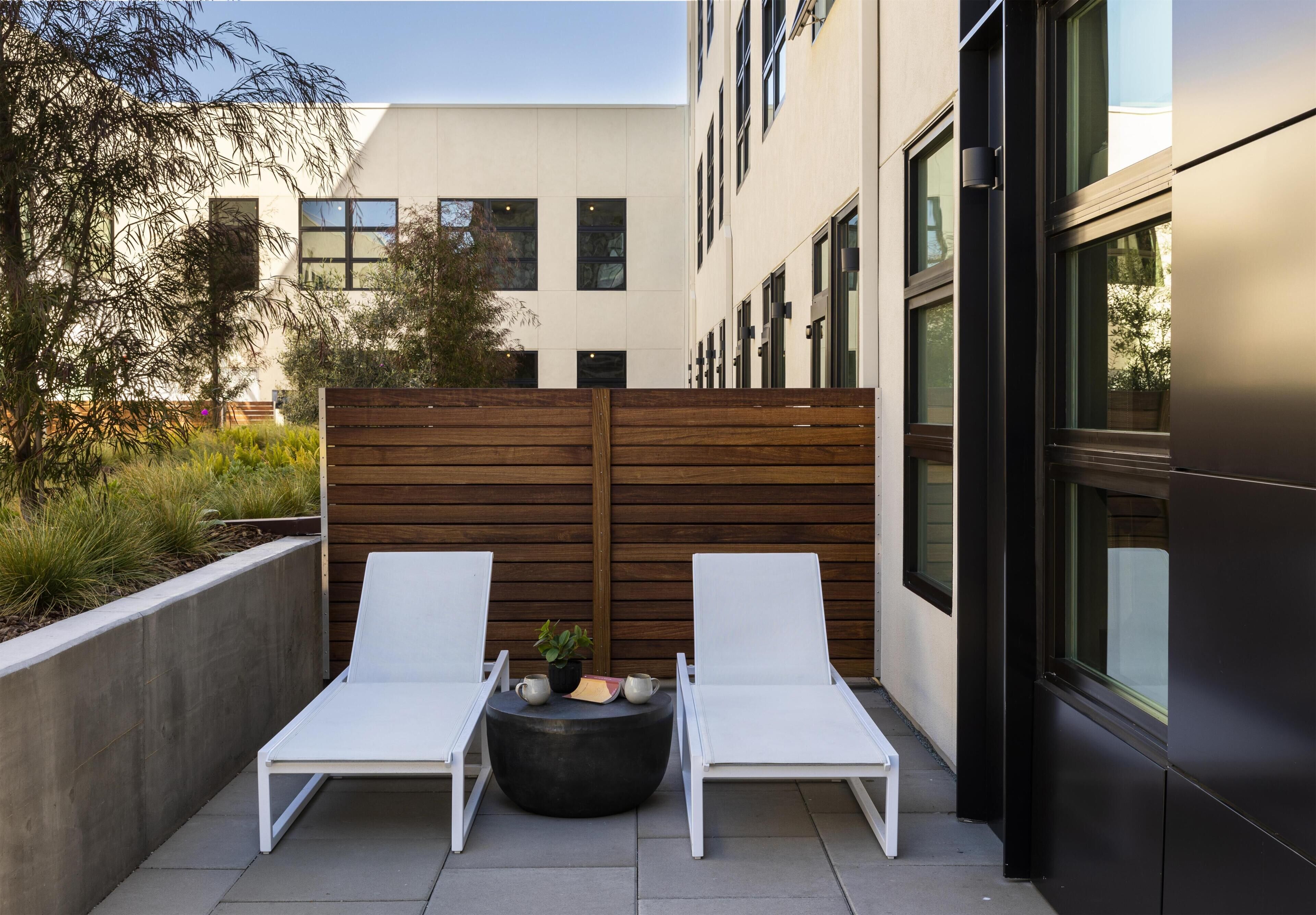 A modern outdoor seating area with two white lounge chairs, a round table with a plant and a book, and a wooden fence.