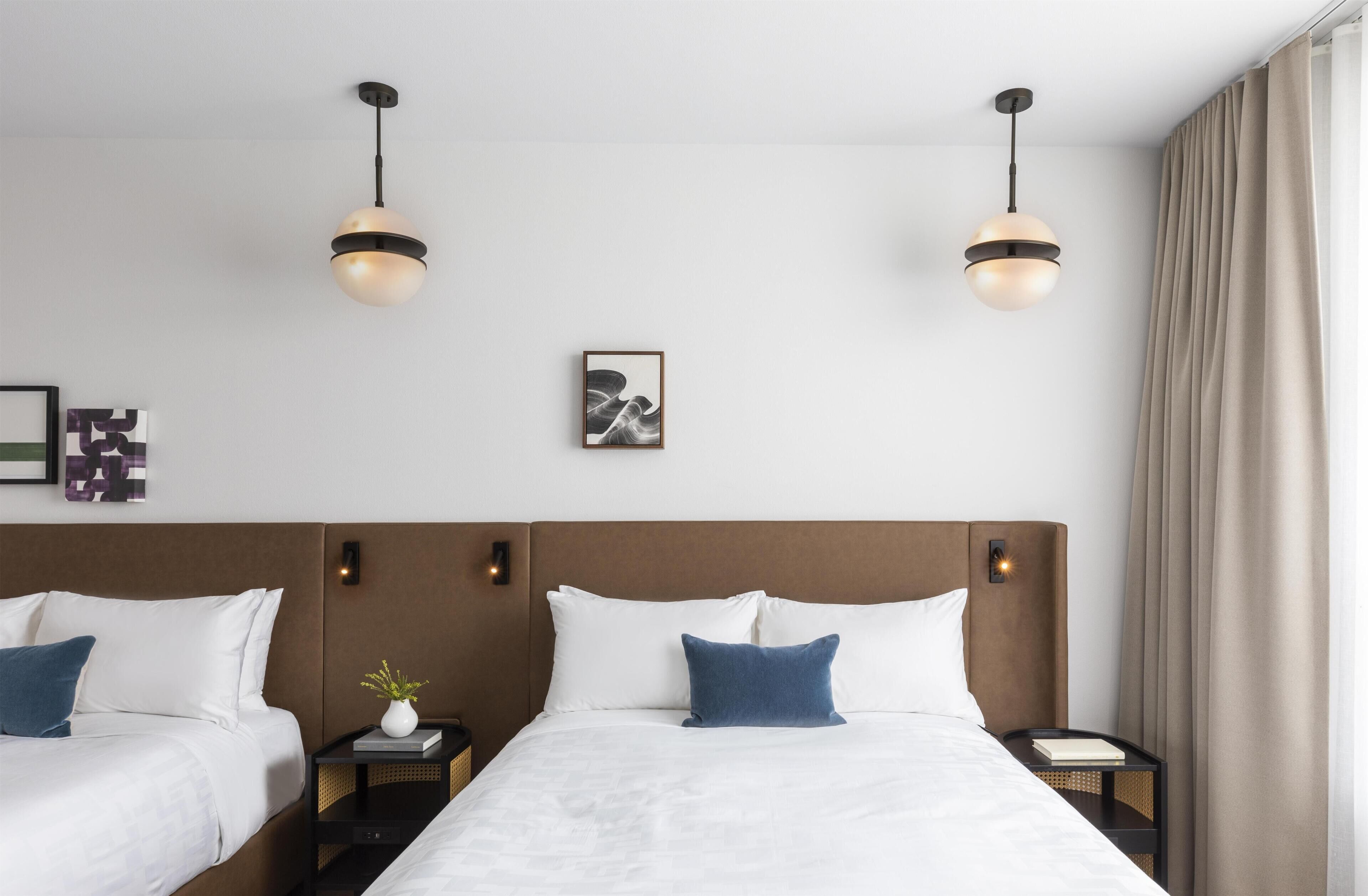 A hotel room with two beds, a large window with curtains, and two pendant lights.