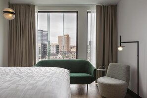 A hotel room with a bed, a green armchair, a grey armchair, a floor lamp, and a view of city buildings through large windows. - Kissel Uptown Oakland, in the Unbound Collection by Hyatt (Oakland)