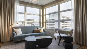 Una sala de estar moderna con sofá, mesa de centro y mesita auxiliar, ventanas amplias con vistas a la ciudad y una lámpara colgante.