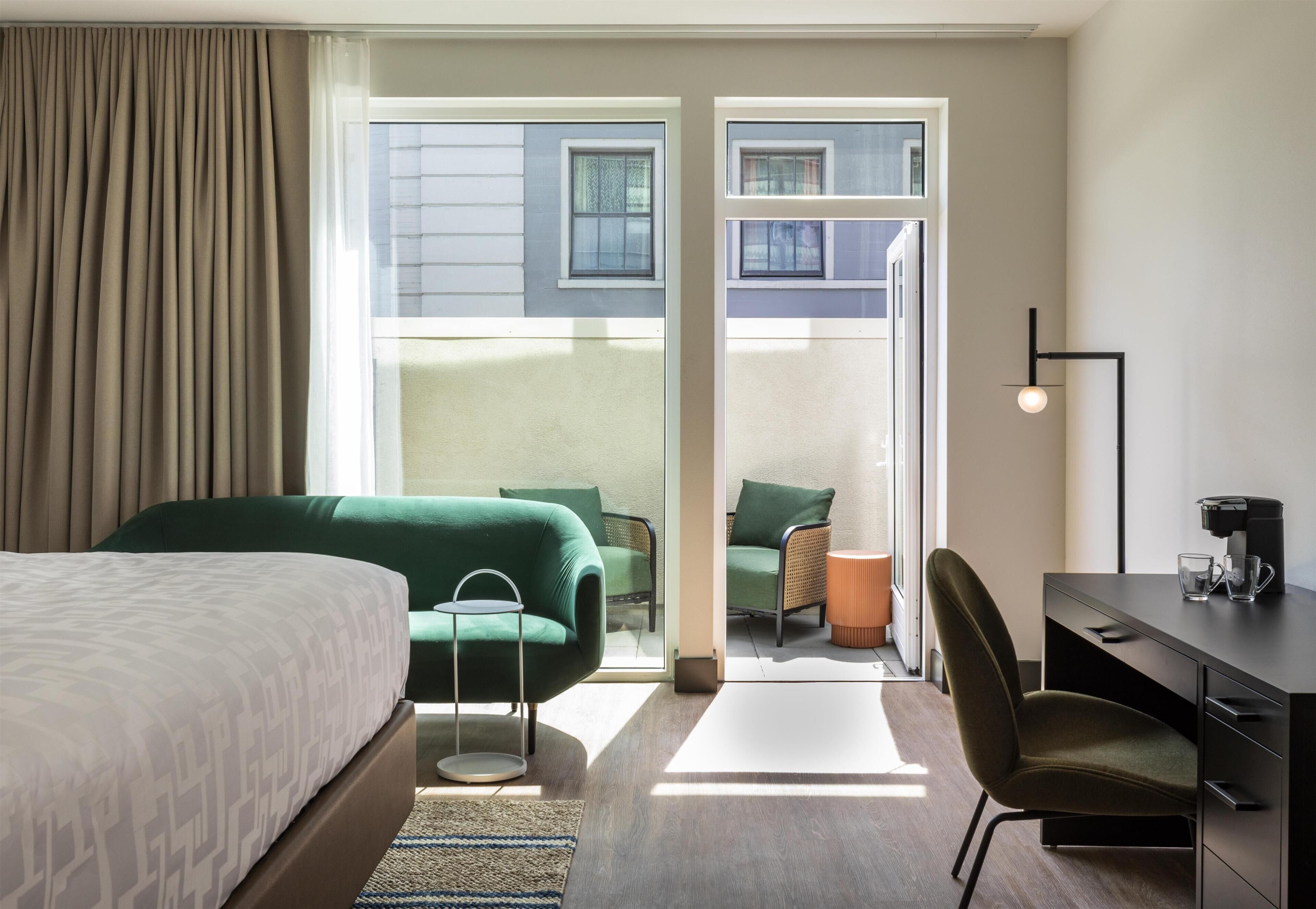 Quarto de hotel com cama, escrivaninha, cadeira, mesinha e vista através de uma porta de vidro.