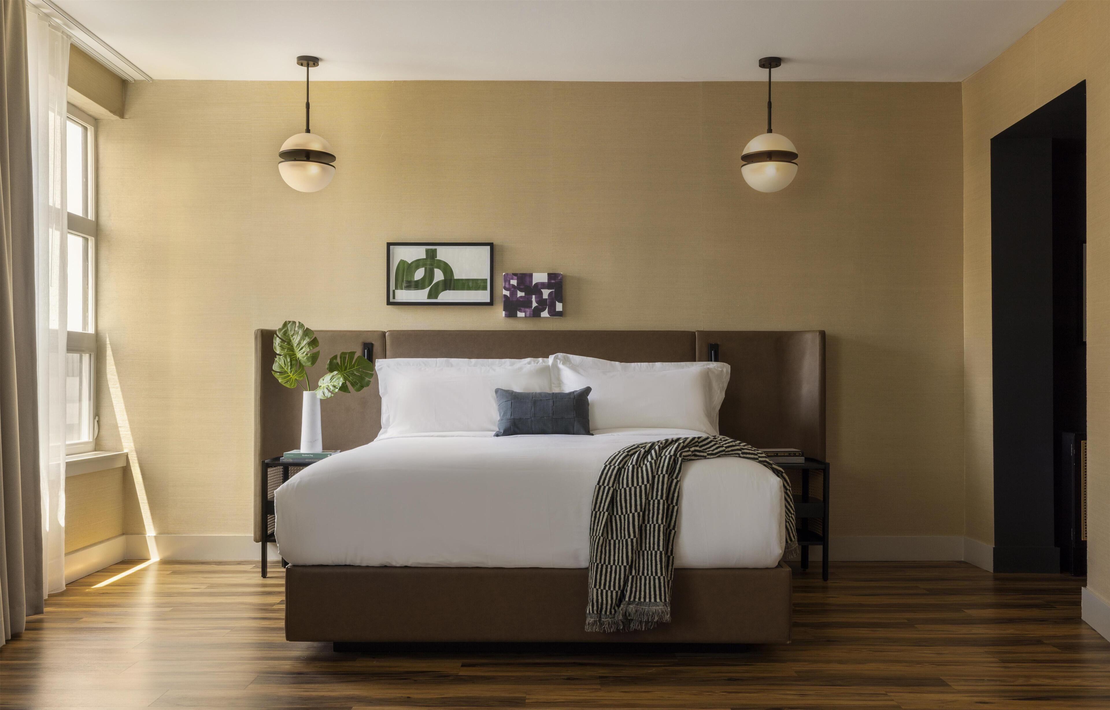 A modern bedroom with a large bed, two pendant lights, a plant, and artwork on the wall.