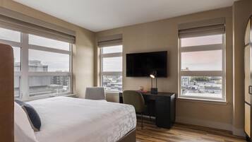 A hotel room with a bed, desk, chair, TV, and large windows offering a city view.