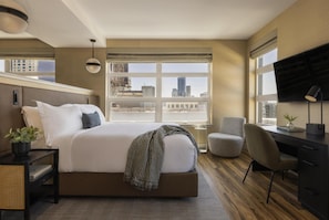 A modern hotel room with a large bed, a desk, a chair, a TV, and a view of the cityscape.