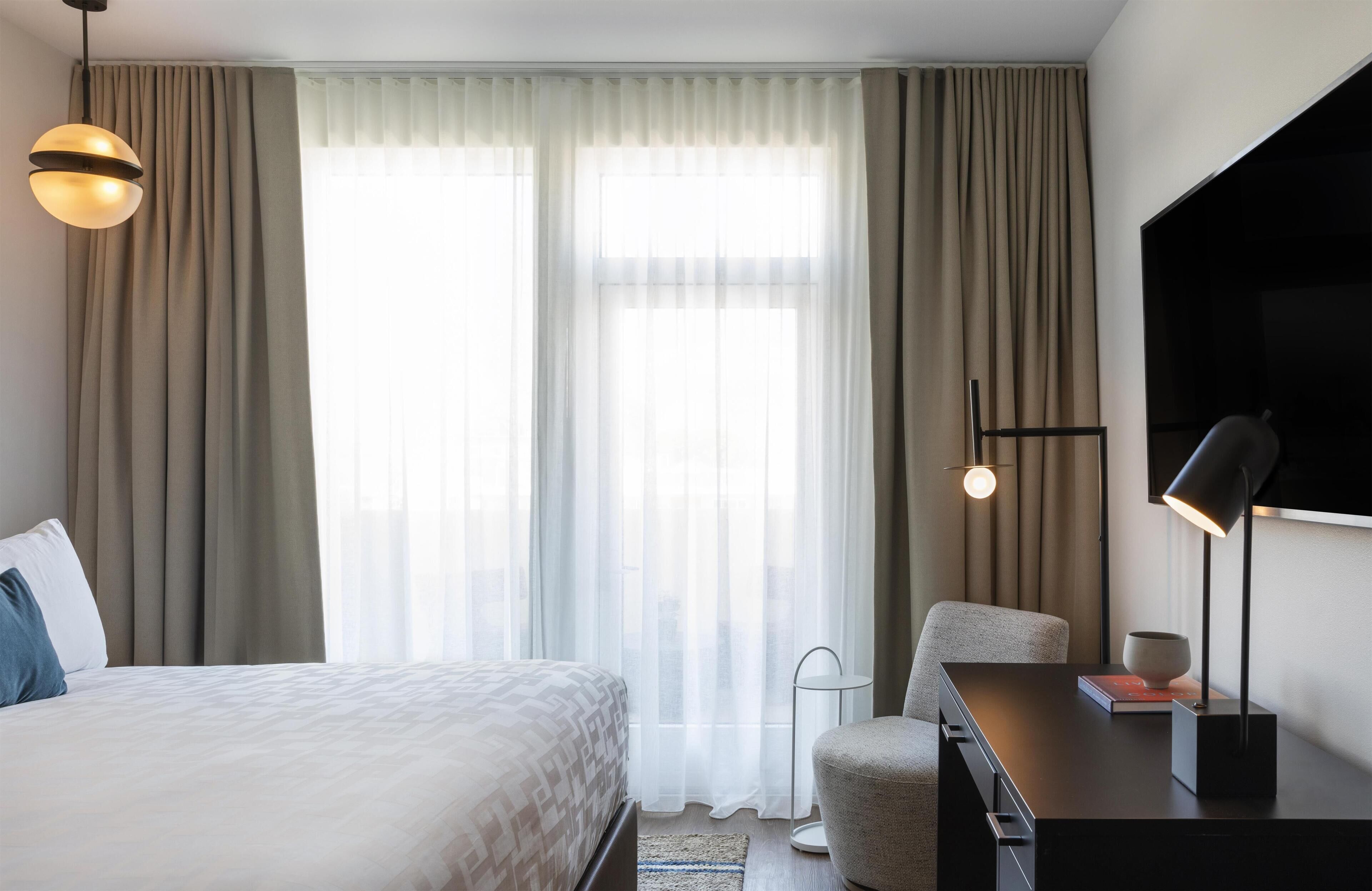A hotel room with a bed, a desk, a chair, a TV, and a window with curtains.