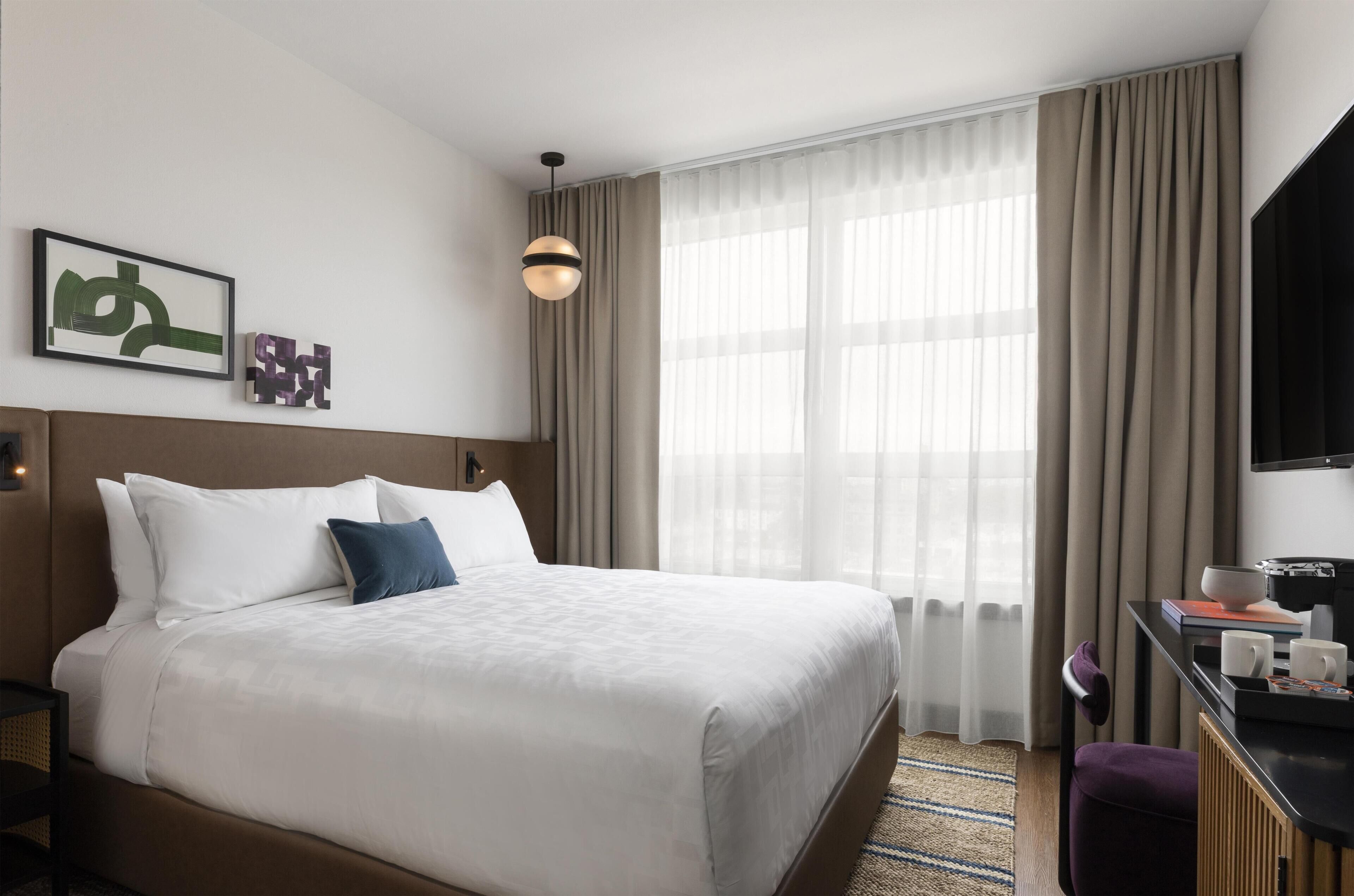 Una habitación de hotel con cama, escritorio con silla, televisor y ventana con cortinas.