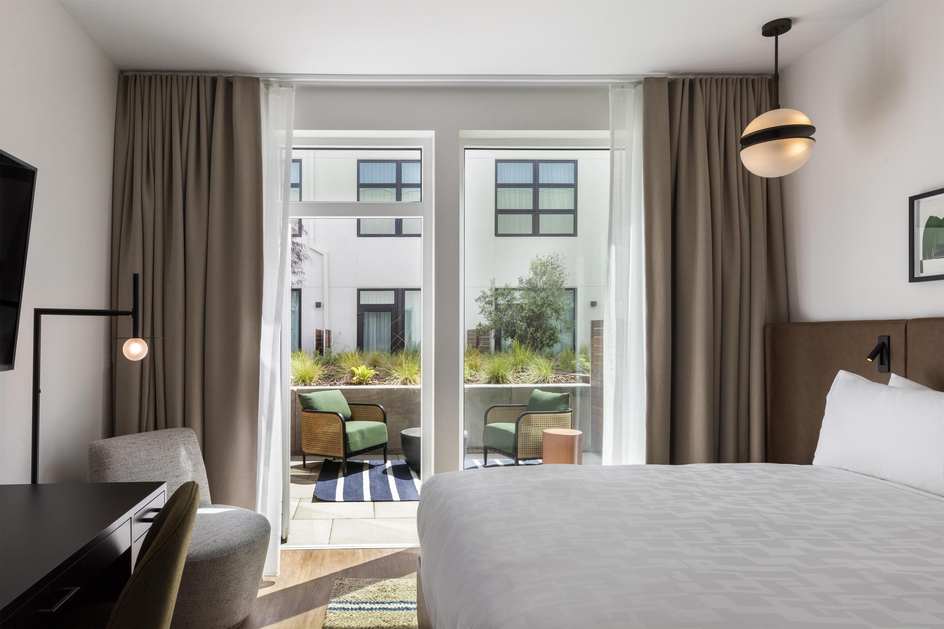 A hotel room with a bed, a desk, a chair, a television, and a view of the outdoor area through a sliding door.