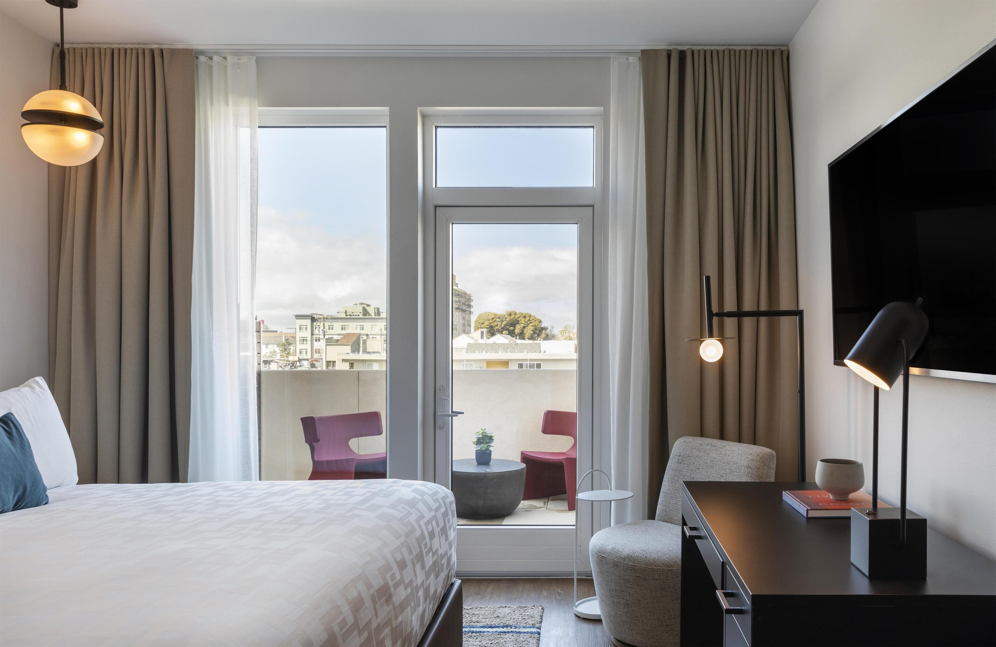 A hotel room with a bed, a desk, a chair, a TV, a balcony with chairs, and curtains.