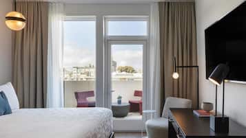 A hotel room with a bed, a desk, a chair, a TV, a balcony with chairs, and curtains.
