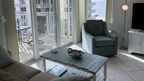 Ein modernes Wohnzimmer mit Sofa, Sessel und Couchtisch. Große Fenster bieten Blick auf einen Balkon und Gebäude.