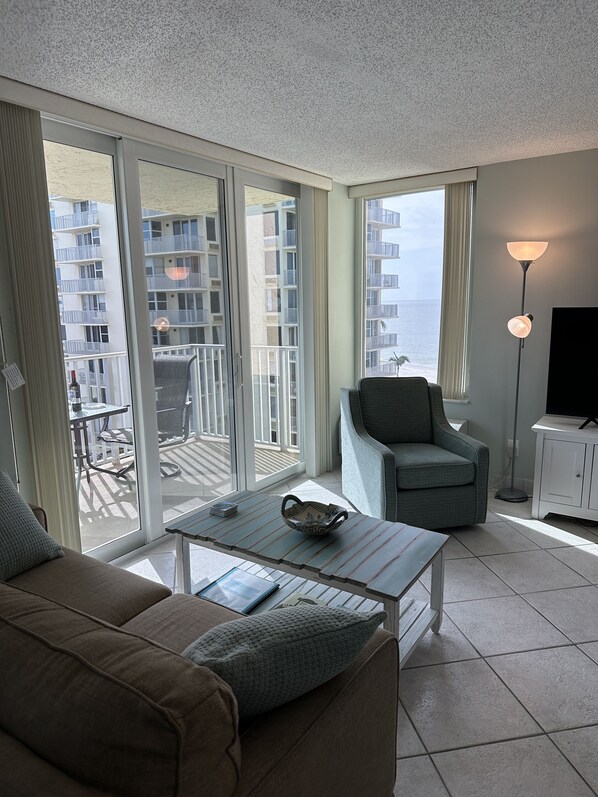 Un salon moderne avec un canapé, un fauteuil et une table basse. De grandes fenêtres offrent une vue sur un balcon et des immeubles.