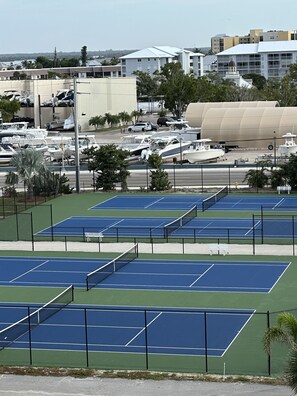 Un complexe de courts de tennis comprenant plusieurs terrains, entouré d’une clôture, et une aire de stationnement où sont garés des bateaux et des véhicules.
