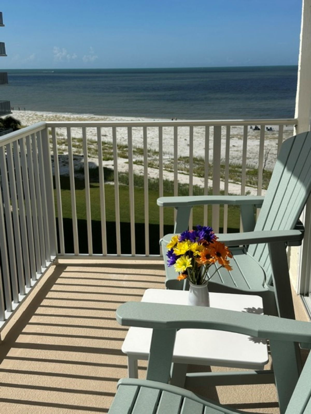 A balcony with a view of the ocean, a white table with a vase of flowers, and two green chairs.