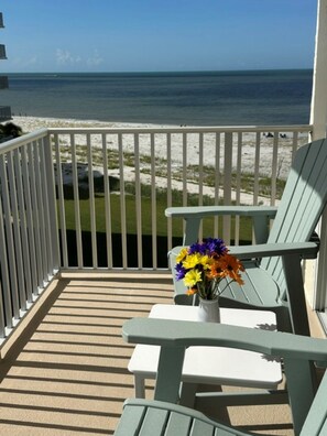 A balcony with a view of the ocean, a white table with a vase of flowers, and two green chairs.