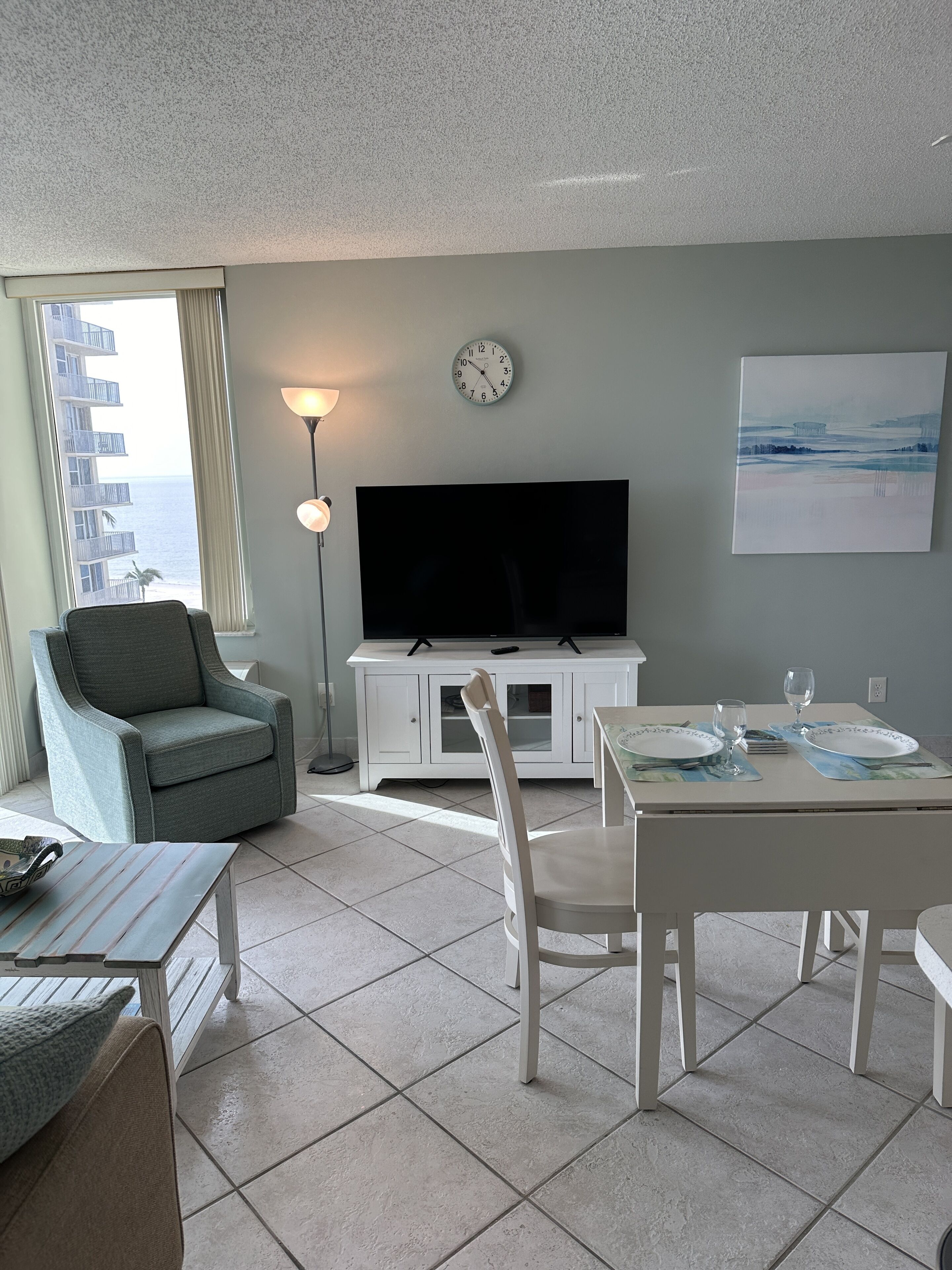 A living room with a TV, dining table, chairs, a sofa, a lamp, and a clock on the wall.