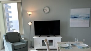 Ein Wohnzimmer mit Fernseher, Esstisch, Stühlen, einer Couch, einer Lampe und einer Wanduhr.