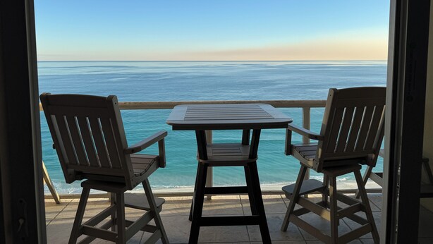A balcony with two wooden chairs and a table, overlooking a calm sea and clear sky. - 🌅 INCREDIBLE View ~ Free Beach Service..SPACIOUS-Remodeled Kitchen~Gulf Front 🏖️ (Navarre)