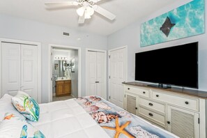 Un dormitorio con una cama, televisión, ventilador de techo y un baño visible a través de una puerta abierta.