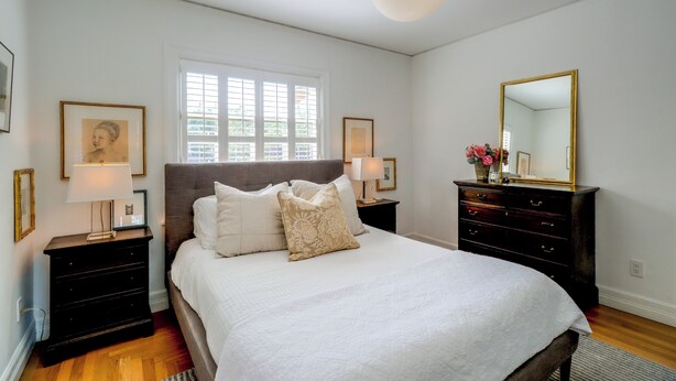 Une chambre à coucher avec un grand lit, deux tables de chevet, une commode, une fenêtre avec des stores et un luminaire suspendu.