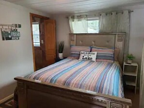 A bedroom with a bed, a sign on the bed, a window with curtains, a wooden door, and a small table with a plant.