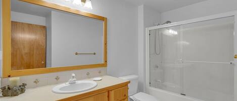 Un baño con tocador de madera, un espejo grande, un lavabo blanco, inodoro y cabina de ducha con paredes de vidrio.